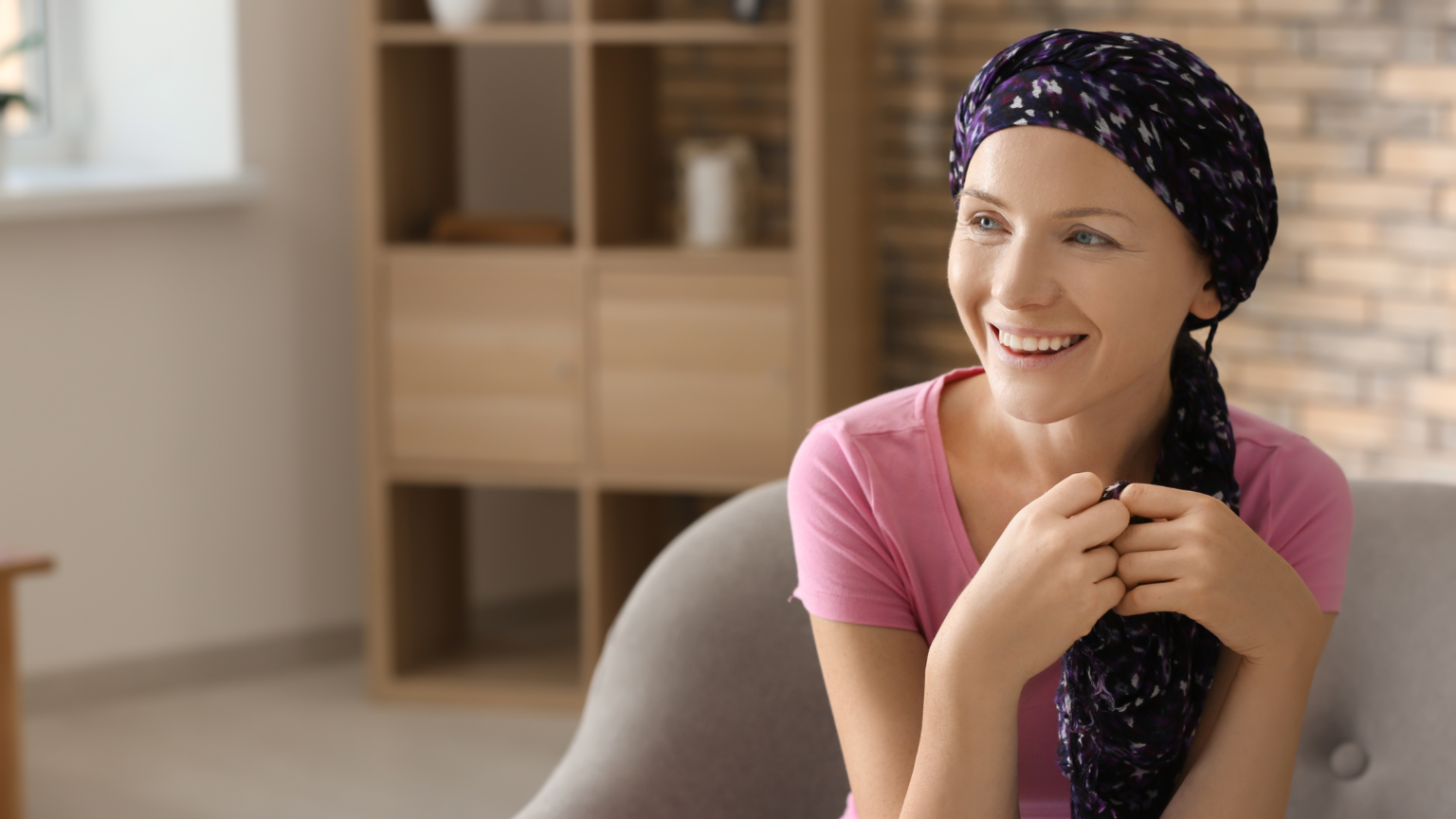in foto una donna sorridente con un turbante sulla testa, seduta su una poltrona: rappresenta una paziente oncologica, che può usufruire dell'estetica oncologica per stare meglio