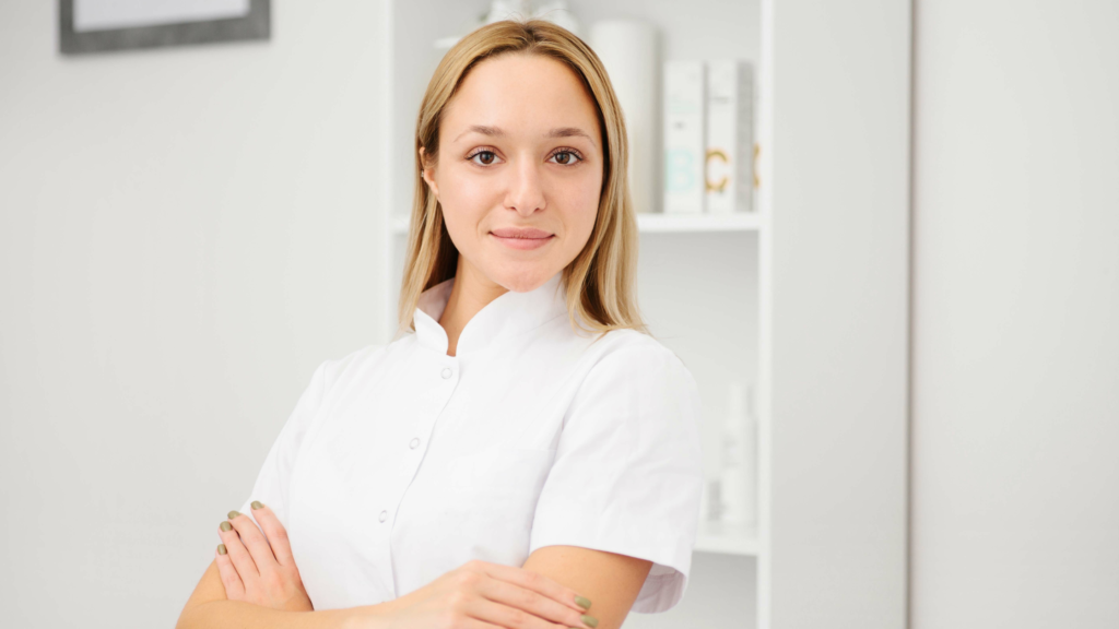 L'immagine ritrae un'estetista professionale con un aspetto curato e un sorriso rassicurante. Indossa una divisa bianca, simbolo di pulizia e professionalità, e ha le braccia incrociate, trasmettendo sicurezza e competenza. Lo sfondo minimalista con scaffali ordinati e prodotti cosmetici suggerisce un ambiente di lavoro moderno e accogliente, ideale per trattamenti estetici e consulenze di bellezza.