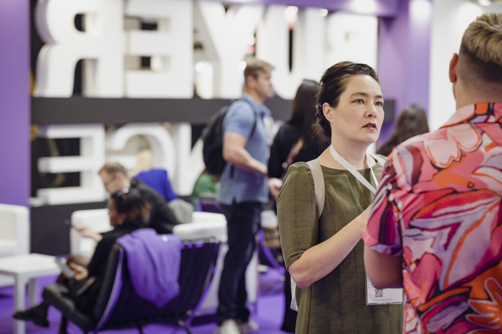 Persone che interagiscono durante Cosmoprof 2025 in un’area dedicata al networking. Una donna in primo piano conversa, mentre sullo sfondo altre persone lavorano e si scambiano idee in un ambiente moderno con decorazioni viola.