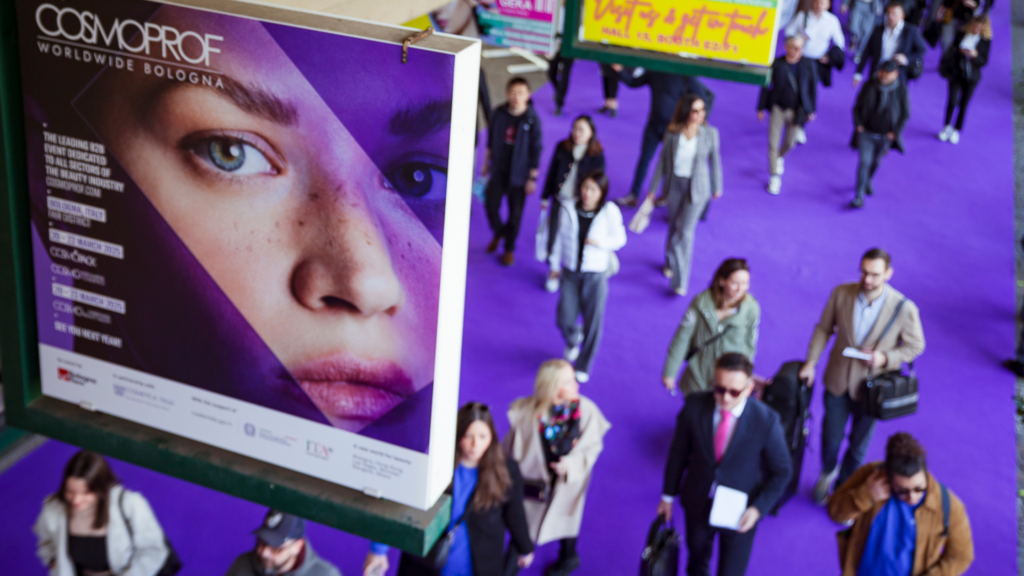 Cartellone pubblicitario di Cosmoprof 2025 con un grande primo piano di un volto femminile su sfondo viola. Sullo sfondo, un corridoio con moquette viola affollato di partecipanti che camminano all’interno dell’evento a Bologna.