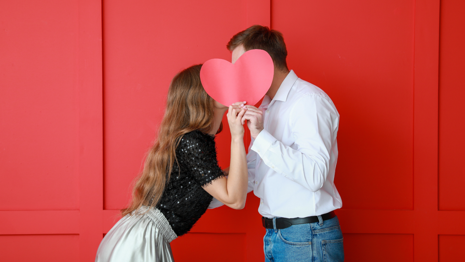 in foto su sfondo rosso, due innamorati che si baciano dietro un cartoncino a cuore. Rappresenta la festività di San valentino 2025.